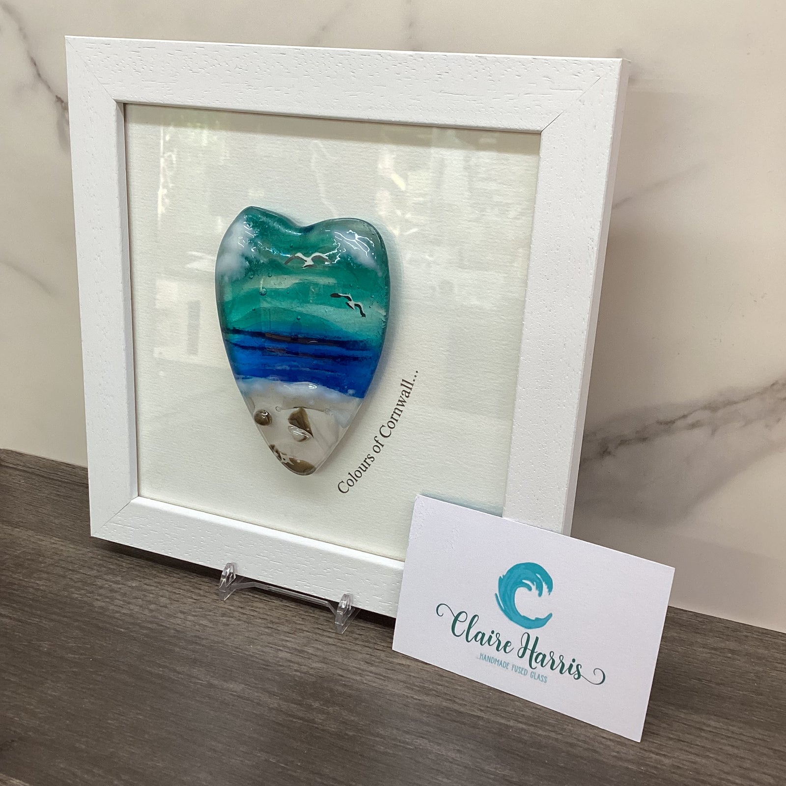 A framed decorative piece featuring a conical shaped sea scene heart with the text 'Colours of Cornwall' up the side. 
