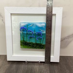 A square white-framed artwork with a colourful embedded scene showing blue water, purple agapanthus flowers, green grass, and small white seagulls, photographed with a metal ruler to demonstrate the height of the piece.