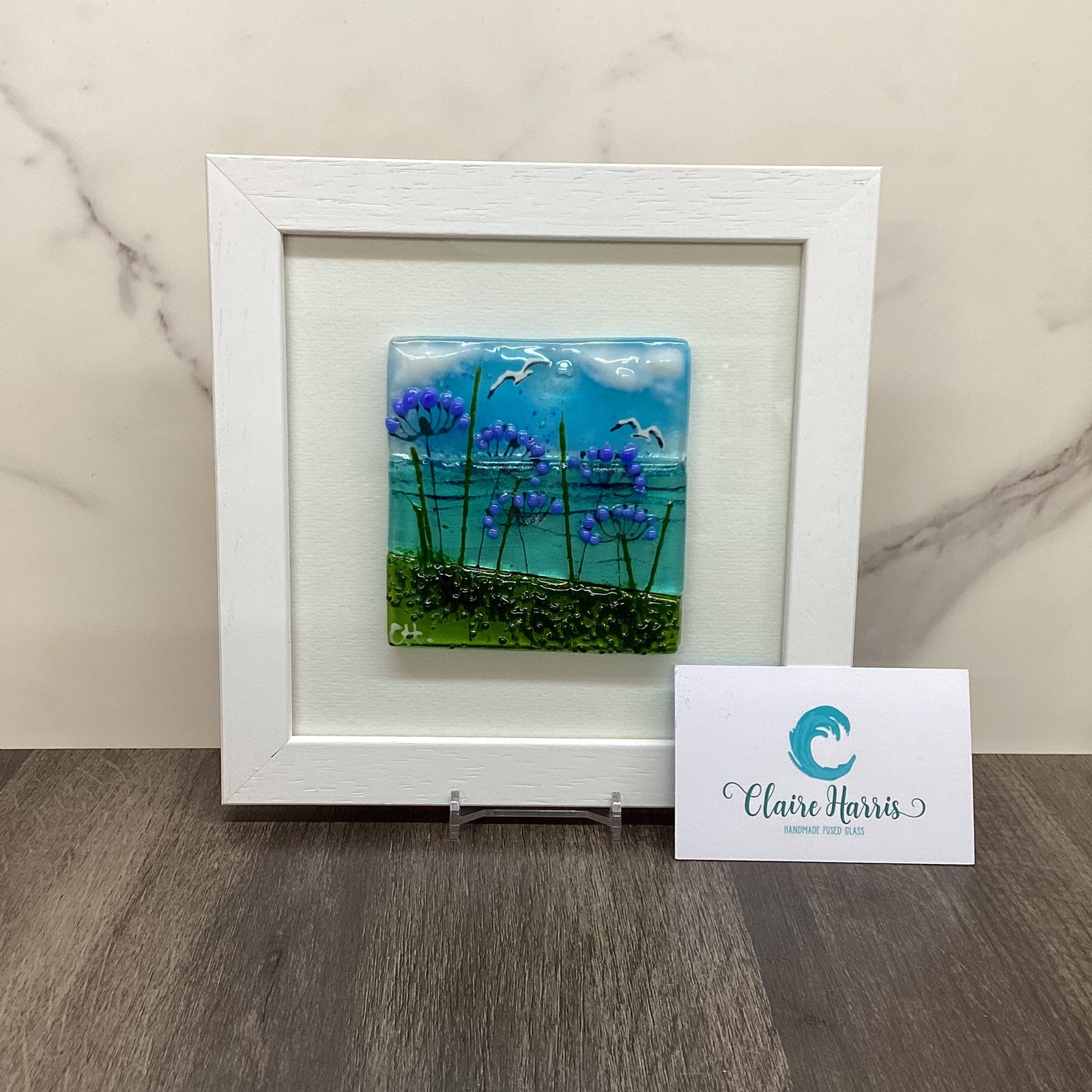 A square white-framed art piece featuring a colourful fused glass landscape with blue sky, purple agapanthus flowers, and green grass, displayed on a marble surface next to a branded business card.