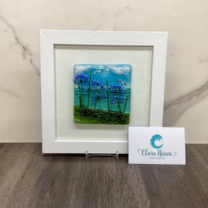 A square white-framed art piece featuring a colourful fused glass landscape with blue sky, purple agapanthus flowers, and green grass, displayed on a marble surface next to a branded business card.