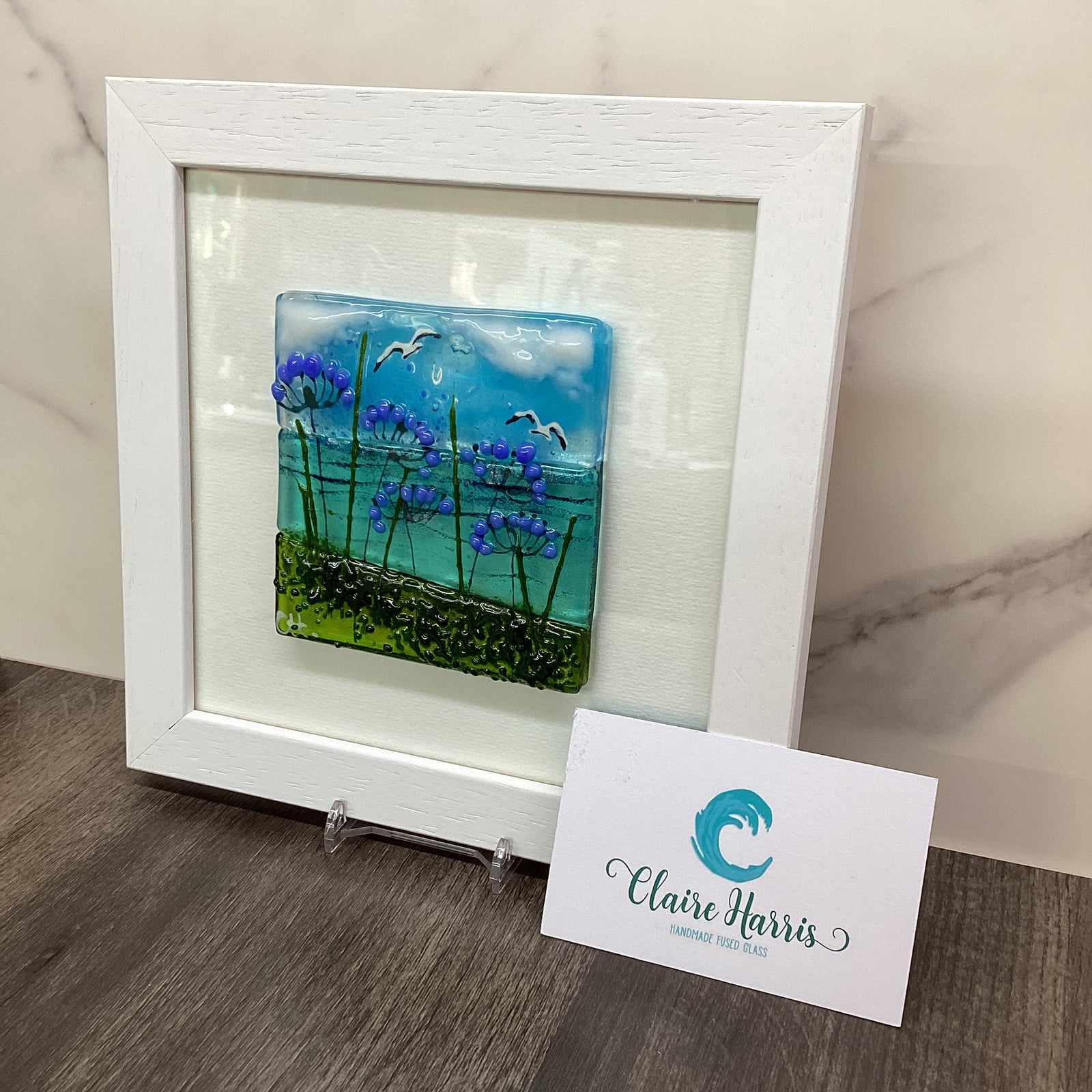 A square white-framed art piece featuring a colourful fused glass landscape with blue sky, purple agapanthus flowers, and green grass, displayed on a marble surface next to a branded business card.