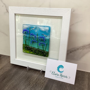 A white square frame containing a colourful fused glass artwork with blue sky, green plants, and purple agapanthus flowers, displayed on a marble surface with a business card attached. View to the right of the product.