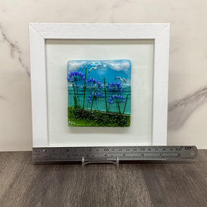 A square white-framed glass art piece featuring blue sky, purple flowers, and green grass, displayed on a marble surface next to a metal ruler for scale.