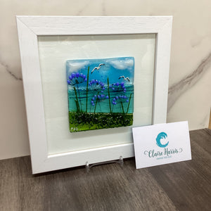 A square white-framed glass artwork featuring a blue and green landscape with purple agapanthus flowers and white birds, displayed on a marble surface with a branded card. Left view of the product.l